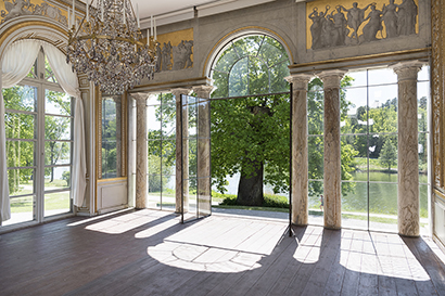 A sunlit hall with large windows overlooking lush trees and water.